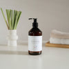 A bottle of hand soap sits on a white table beside a vase filled with fresh lemongrass stalks.
