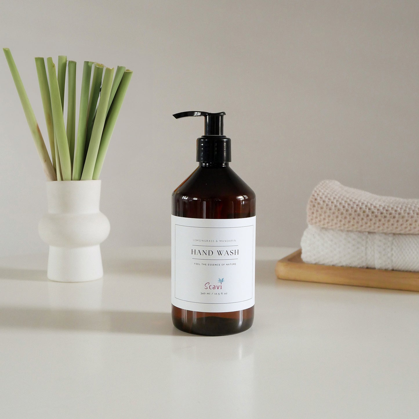 A bottle of hand soap sits on a white table beside a vase filled with fresh lemongrass stalks.